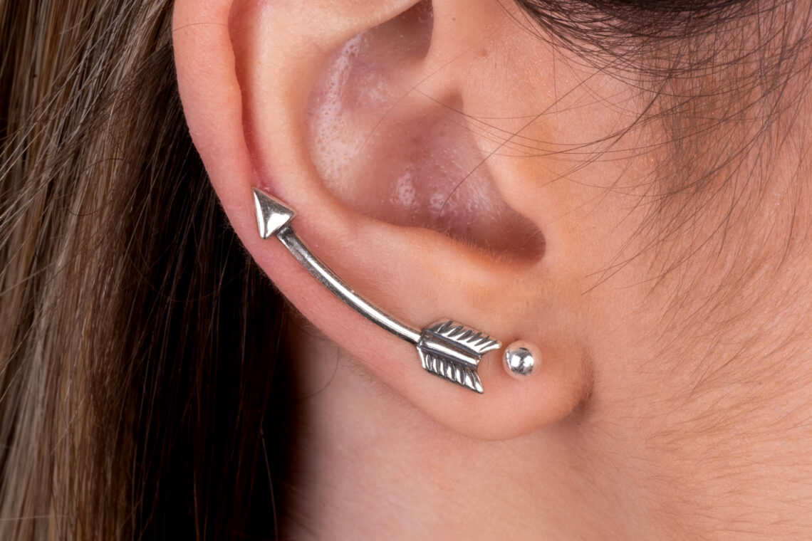 Orelha feminina com dois brincos; No primeiro furo a modelo usa uma bolinha de pérola e no segundo furo uma peça de brinco ear cuff prata no formato de seta.