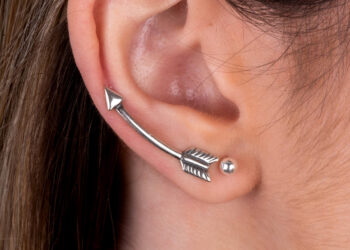 Orelha feminina com dois brincos; No primeiro furo a modelo usa uma bolinha de pérola e no segundo furo uma peça de brinco ear cuff prata no formato de seta.